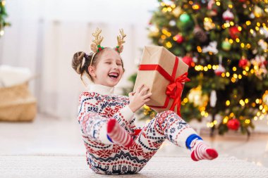 Kırmızı kurdeleli bir kutuyla pijamalı sevimli bir kız. Neşeli ve elinde bir hediye tutuyor. Arka plandaki çelenklerin ışığında Noel ağacı.