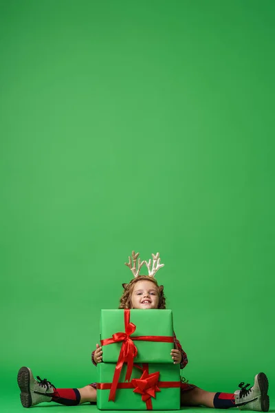 可爱的小女孩 卷曲的头发 金鹿角 坐在工作室绿色背景的礼品盒上 孩子高兴地微笑着 看着镜框 复制空间 — 图库照片