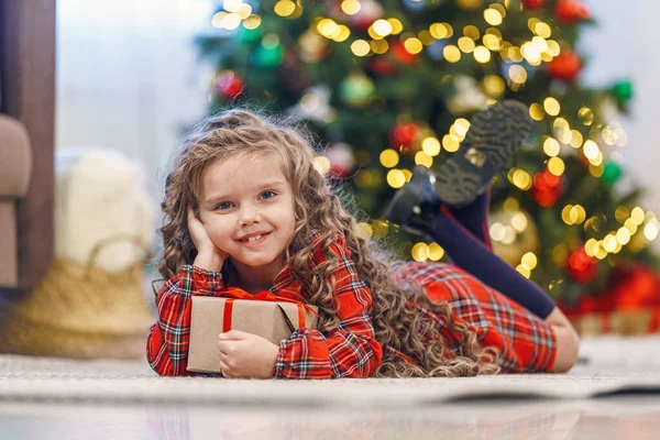 身穿格子花裙 头戴红丝带礼品盒的快乐小女孩躺在地板上 手里拿着礼物 背景中绿油油的圣诞树 — 图库照片