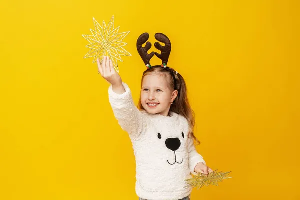 黄色の背景にクリスマスの星と鹿の角を持つ幸せな女の子 子供は幸せで楽しくクリスマスの準備をしています スペースのコピー — ストック写真