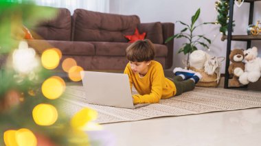 Küçük bir çocuk, sarı kazaklı, karnının üstünde yatıyor, gülümsüyor ve dizüstü bilgisayar ekranına bakıyor. Arka planda, Noel süslemeleri ve parıldayan ışıklar..