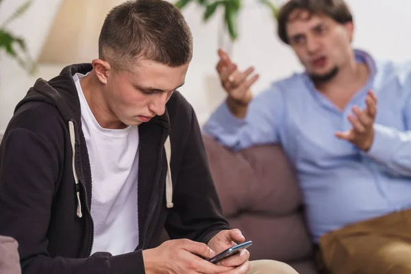 indifferent difficult teenager is sitting on couch with smartphone on Internet. father screams and threatens his son. Misunderstanding between father and son. Internet addiction and gadget addiction