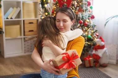 Ren geyiği boynuzları takmış sevgi dolu anne ve kızı arka plandaki Noel ağacına sarılıyor. Kafasında geyik boynuzları olan küçük kız mutlu ve gülüyor. Sarılmak, elinde Noel hediyesi kutusu tutmak.
