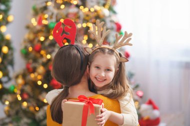 Ren geyiği boynuzları takmış sevgi dolu anne ve kızı arka plandaki Noel ağacına sarılıyor. Kafasında geyik boynuzları olan küçük kız mutlu ve gülüyor. Sarılmak, elinde Noel hediyesi kutusu tutmak.