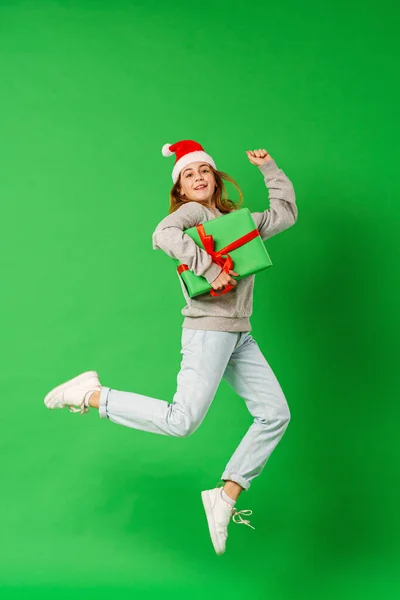 一个快乐的微笑的女孩 手里拿着红丝带礼品盒 头上戴着一顶圣诞帽 在绿色的背景上跳跃着 准备圣诞节打折销售动态图像 — 图库照片