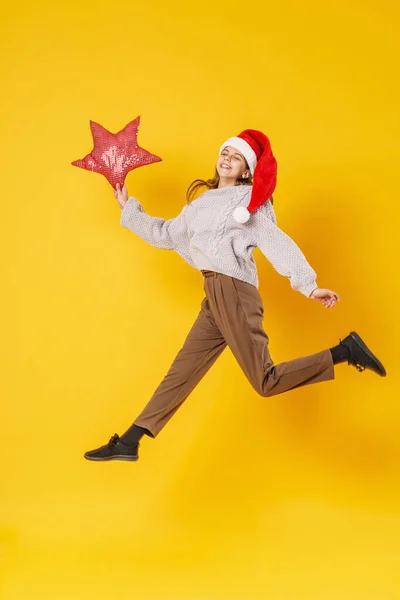 一个快乐的微笑的女孩 手里拿着一颗红星 头上戴着一顶圣诞老人的帽子 在黄色的背景上跳跃着 准备圣诞节打折销售动态图像 — 图库照片