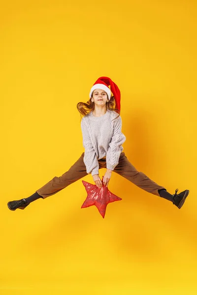 一个快乐的微笑的女孩 手里拿着一颗红星 头上戴着一顶圣诞老人的帽子 在黄色的背景上跳跃着 准备圣诞节打折销售动态图像 — 图库照片