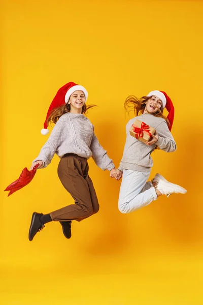 两个笑容满面的女朋友 手里拿着红星 手里拿着礼物 头上顶着一顶圣诞老人的帽子 在黄色的背景下跳了起来 准备圣诞节打折销售动态图像 — 图库照片
