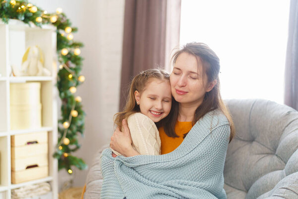 С Днем Матери. Маленькая дочь поздравляет свою мать и прижимается к ней, завернутая в одеяло, сидя на заднем плане огней. Мама и девочка улыбаются и обнимаются. Семейный отдых и единство.