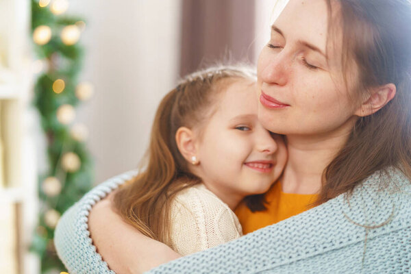 С Днем Матери. Маленькая дочь поздравляет свою мать и прижимается к ней, завернувшись в одеяло, сидя дома на фоне огней. Мама и девочка улыбаются и обнимаются. Крупный план