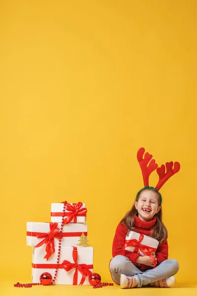 可爱的小女孩5岁 戴着红鹿角 坐在工作室黄色背景的礼品盒上 孩子高兴地微笑着 看着镜框 复制空间 — 图库照片