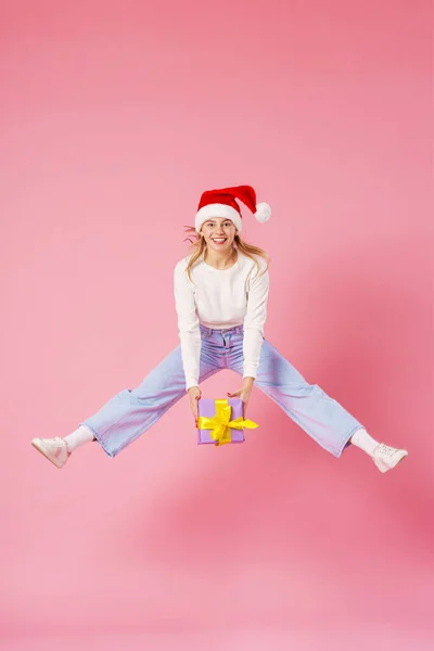 快乐的微笑着的女孩 手里拿着红丝带礼品盒 头上戴着一顶圣诞老人帽 背景是粉色的 准备圣诞节打折销售动态图像 — 图库照片