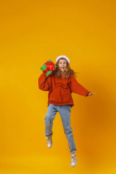 一个快乐的微笑的小女孩 手里拿着礼品盒 头上顶着一顶圣诞老人的帽子 在黄色的背景上跳跃着 准备圣诞节打折销售动态图像 — 图库照片