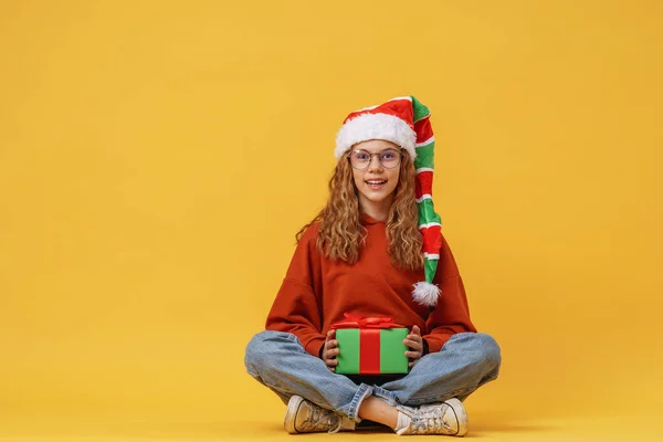 在工作室里 戴着圣诞老人侏儒帽的小女孩拿着一个圣诞礼品盒坐在黄色的背景上 孩子们笑着收到了许多圣诞礼物 — 图库照片