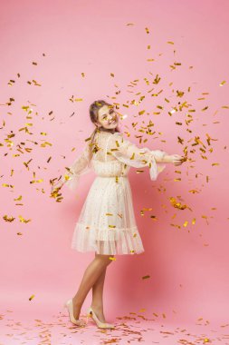Mutlu noeller. Açık renk elbiseli, dalgalı saçları olan güzel çekici bir kız, altın renginde düşüşün altında dans ediyor, stüdyoda pembe bir arka planda uçan konfeti. Büyürken. Boşluğu kopyala