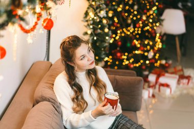 Çekici bir kız, rahat bir odada, elinde kupayla, beyaz örülü bir kazakla bir kanepede oturuyor. Güzellik gülümsemeleri, bakışları, evin içi Noel için süslenmişti. kör edici ışık.
