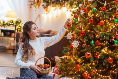 Mutlu Noeller ve mutlu yıllar. Beyaz kazaklı güzel bir genç kız bir Noel ağacının yanında oturuyor ve süsleyerek süslenmiş bir evde kırmızı bir balon asıyor.