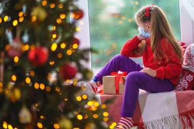 Sevimli küçük kız elinde hediye kutusuyla pencerenin kenarında oturuyor. Dekore edilmiş Noel ağacı, battaniyeli ve penceresinin yanında yastıklı çocuk. Çocuğun yüzünde maske. Mutsuz çocuk karantinada.