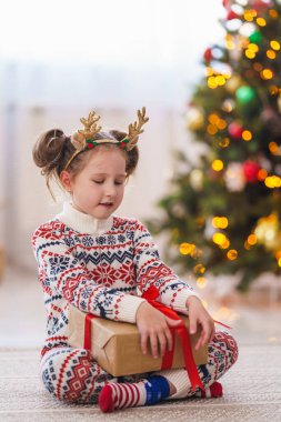 Kırmızı kurdeleli bir kutuyla pijamalı sevimli bir kız. Neşeli ve elinde bir hediye tutuyor. Arka plandaki çelenklerin ışığında Noel ağacı.