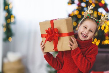 Kırmızı kazaklı, kırmızı kurdeleli şirin, neşeli bir kız. Neşeli ve elinde bir hediye tutuyor. Arka plandaki çelenklerin ışığında Noel ağacı.