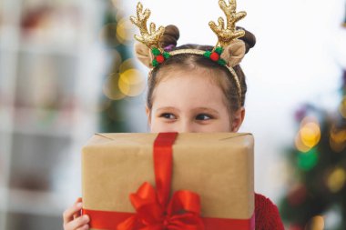 Kırmızı kazaklı, kırmızı kurdeleli şirin, neşeli bir kız. Neşeli ve elinde bir hediye tutuyor. Arka plandaki çelenklerin ışığında Noel ağacı.