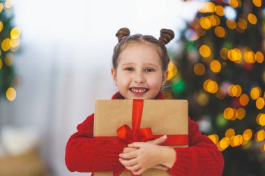 Elinde bir kutuyla kırmızı kurdeleyle bağlanmış sevimli, neşeli küçük bir kız. Hediye kucaklamaları vücuda baskı yapıyor. Arka plandaki çelenklerin ışığında Noel ağacı.