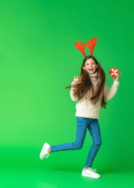 一个快乐快乐的女孩穿着针织毛衣 手里拿着一个盒子的大鹿角作为圣诞礼物的画像 一个孩子在绿色的背景上跑着跳着 动态图像 复制空间 — 图库照片