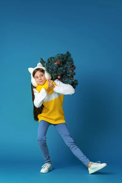 身穿针织毛衣 头戴毛皮耳机 肩上挂着红色圣诞彩球的快乐少女 穿着蓝色背景的衣服走路 动态图像 — 图库照片