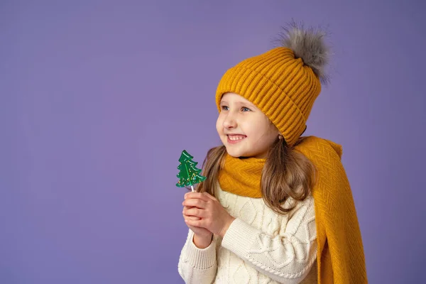 黄色の帽子とスカーフでブロンドの髪で5歳の小さなヨーロッパの女の子 彼女の手の中に緑のクリスマスツリーの形でキャンディーを保持 紫の背景に喜んで笑顔立っています — ストック写真