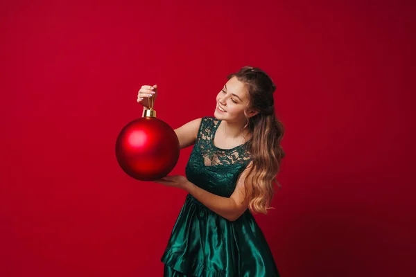 在工作室里 穿着绿色衣服的漂亮可爱的女孩站在一个红色背景的大大的红色圣诞树球旁边 一个少女高兴地微笑着 看着球 复制空间 — 图库照片