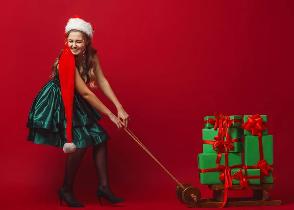 穿着绿色衣服头戴圣诞帽的年轻貌美的姑娘 骑着雪橇 送上了新年礼物 孤零零的红色背景 圣诞节大甩卖 复制空间 全面增长 — 图库照片