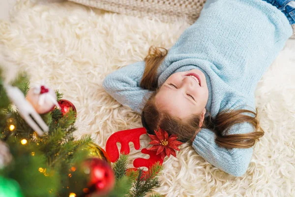 Mavi kazaklı ve kırmızı geyik boynuzlu küçük kız yerde bir hediye kutusuyla bayram süslenmiş bir Noel ağacının yanında yatıyor. Çocuk evde bir gülümseme ve neşeyle kameraya bakar.