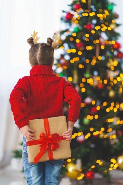 Kırmızı kazaklı, kırmızı kurdeleli şirin, neşeli bir kız. Arkasında bir armağan tutuyordu. Arka plandaki çelenklerin ışığında Noel ağacı.