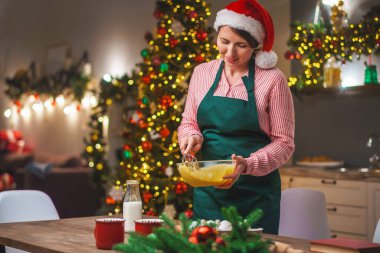 Bir kadın Noel arifesinde mutfakta yemek hazırlar. Önlüklü bir kız hamurun malzemelerini bir kasede karıştırıyor. Mutlu Noeller ve mutlu Noeller. İçi Noel ışıklarıyla süslenmiş..