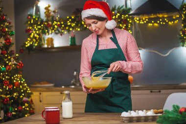 Bir kadın Noel arifesinde mutfakta yemek hazırlar. Önlüklü bir kız hamurun malzemelerini bir kasede karıştırıyor. Mutlu Noeller ve mutlu Noeller. İçi Noel ışıklarıyla süslenmiş..