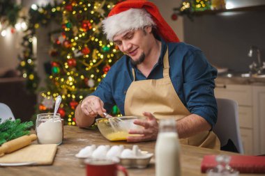 Bir adam Noel arifesinde mutfakta yemek hazırlar. Önlük ve Noel Baba şapkası takmış genç bir adam. Mutlu Noeller ve mutlu Noeller. Noel ışıklarıyla donatılmış..