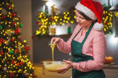 Bir kadın Noel arifesinde mutfakta yemek hazırlar. Önlüklü bir kız hamurun malzemelerini bir kasede karıştırıyor. Mutlu Noeller ve mutlu Noeller. İçi Noel ışıklarıyla süslenmiş..