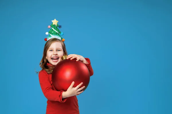 快乐的小女孩戴着装饰过的头巾 带着圣诞红气球 蓝色背景在工作室里 头戴圣诞树的孩子高兴地笑着 并收到圣诞礼物 — 图库照片