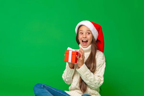 快乐快乐的未成年少女 戴着一顶绿色背景的圣诞老人帽 一个拿着礼品盒的有趣的孩子很高兴收到礼物 送礼物新年销售 — 图库照片