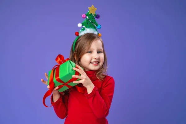快乐的小女孩戴着装饰过的头巾 在工作室里拿着紫色背景的圣诞红气球 头戴圣诞树的孩子高兴地笑着 并收到圣诞礼物 — 图库照片