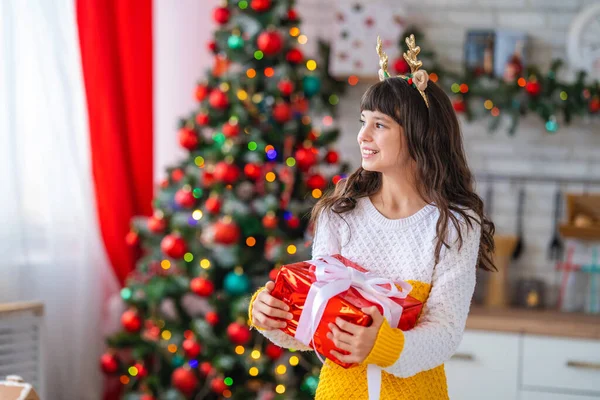 一个甜美的 快乐的 十几岁的女孩 手里拿着一个盒子 用带子捆着 那孩子笑了 手里拿着礼物 背景中绿油油的圣诞树 — 图库照片