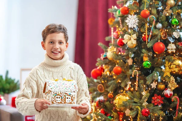 圣诞快乐 一个穿着针织毛衣 拿着礼品盒的快乐男孩站在一棵装饰着闪烁灯光的圣诞树旁边 孩子对他的礼物喜形于色 — 图库照片