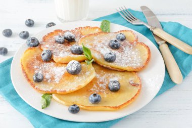 Böğürtlenli krep ve pudra şekeri, mavi peçete, çatal, bir bardak süt, bıçak, üst manzara.