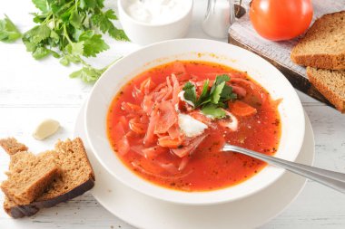 Kırmızı çorba, ekşi kremalı borş ve beyaz tabakta otlar, bir kaşık, beyaz ahşap masada ekmek.