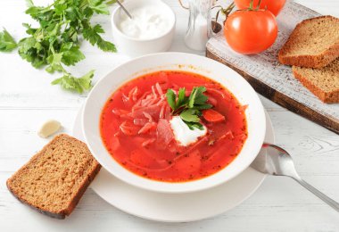 Ekşi kremalı kırmızı borş ve beyaz bir tabakta otlar, bir kaşık, beyaz ahşap bir masada ekmek