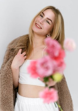 Kahverengi kürklü güzel beyaz sarışın kadın elinde bir buket pembe çiçek ve beyaz bir arka planda gülümseme tutuyor.