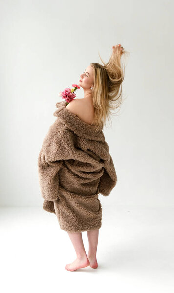 beautiful white young blonde woman in brown furry coat holds a bouquet of pink flowers and raises her hand on a white background full-length