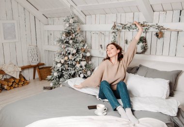 Beyaz tenli, güzel, kot pantolonlu, bej kazaklı bir kız elinde kahve fincanı, elinde Noel ağacı koluyla yatağın lambasında oturuyor.