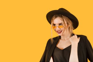 Siyah ceketli, güneş gözlüklü ve sarı arka planda gülümseyen genç beyaz kadın.