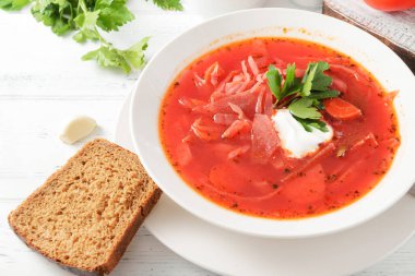 Ekşi kremalı kırmızı borş ve beyaz tabakta otlar, beyaz ahşap bir masada ekmek.